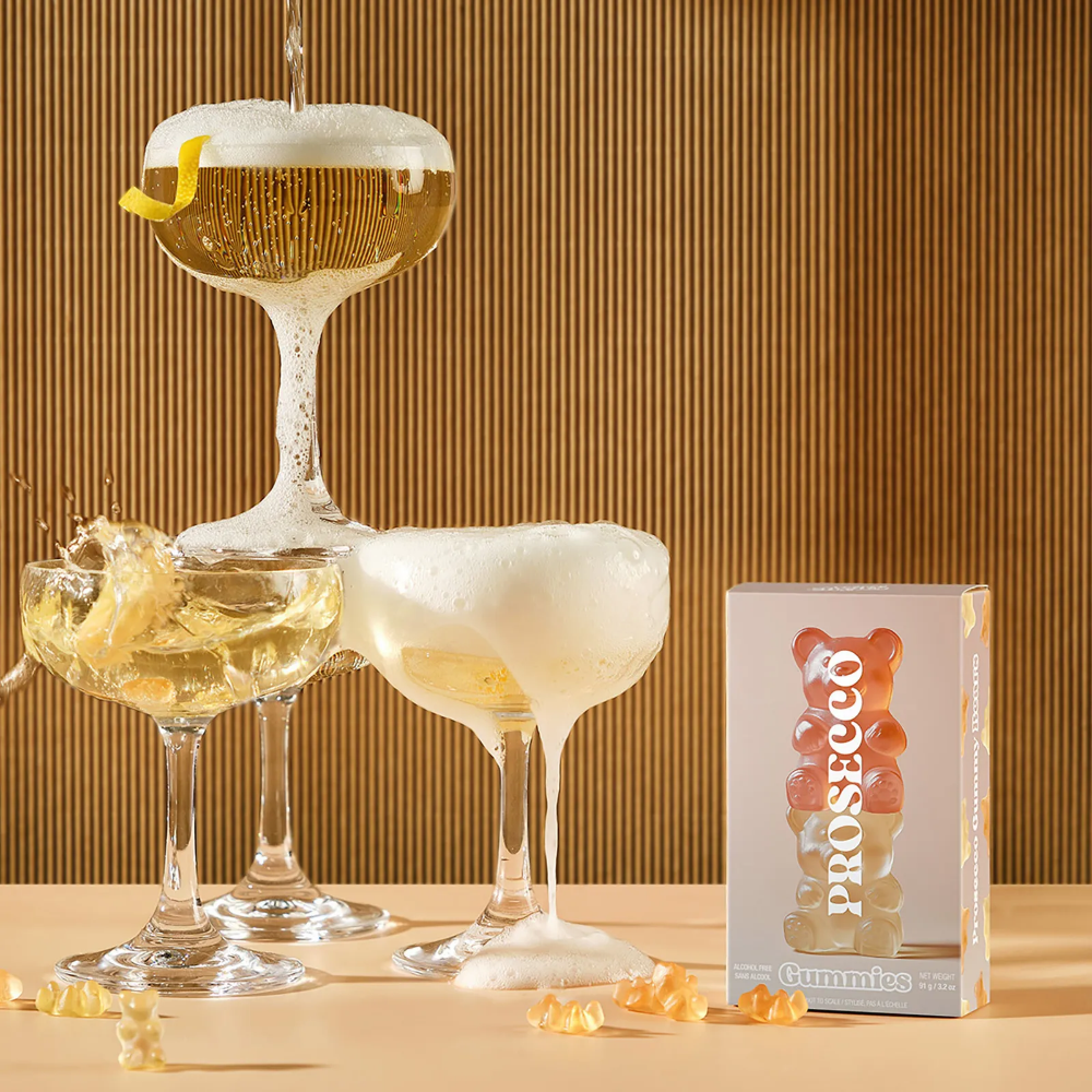 Vesper Bites Prosecco Bears gummies 100g in a blush pink box with a stack of orange and gold gummy bears printed on the front, surrounded by loose gummy bears and glasses.