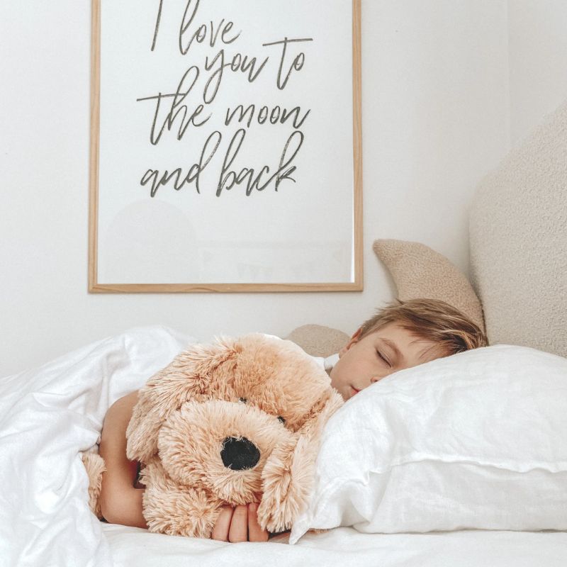 Toddler cuddling a soft tan weighted puppy plush toy while resting on a bed.