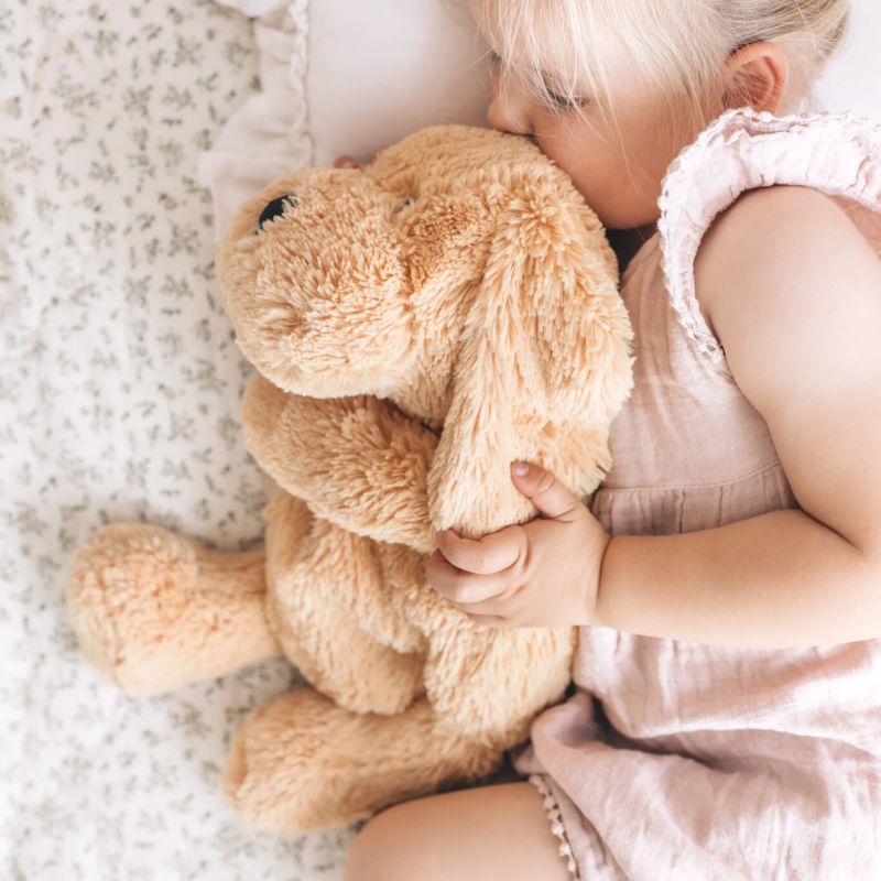 Toddler cuddling a soft tan weighted puppy plush toy while resting on a bed.
