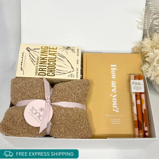 A warming heat pillow, Grounded pleasures drinking chocolate, Fox and Fallow 3 pack of ballpoint pens and 30 day check in activity journal to help with mood and mindfulness all gift wrapped in a beautiful gift box by Wishing You Well