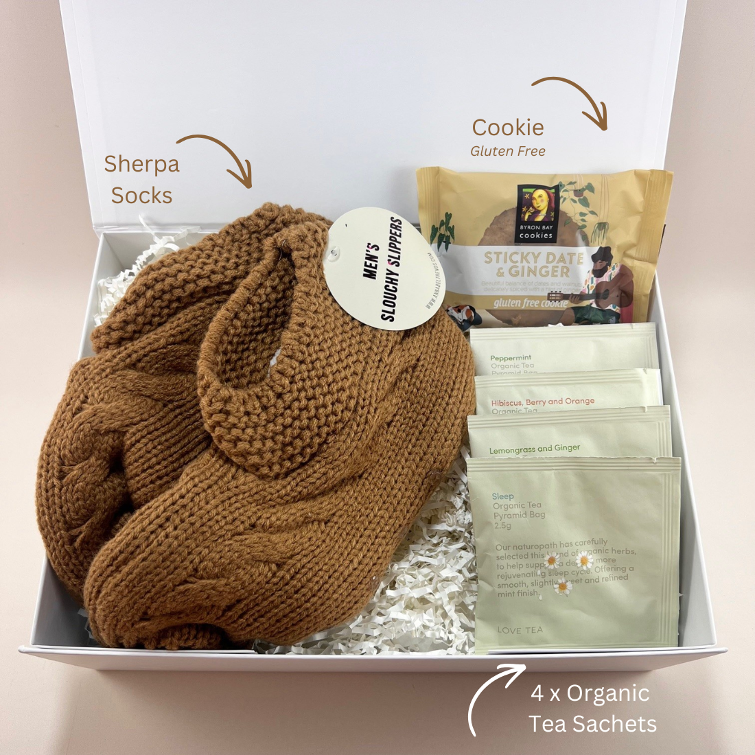 Flat lay of Mini Comforts Care Package contents — men's sherpa socks, four organic tea sachets, and a Byron Bay Cookies sticky date cookie in a white gift box with labels