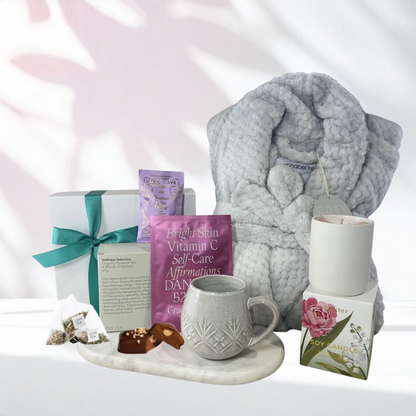 Cosy Self-Care Retreat gift box with a grey plush robe, organic wellness tea selection, textured stoneware mug, soy candle, chocolate pieces and face mask displayed on a marble tray