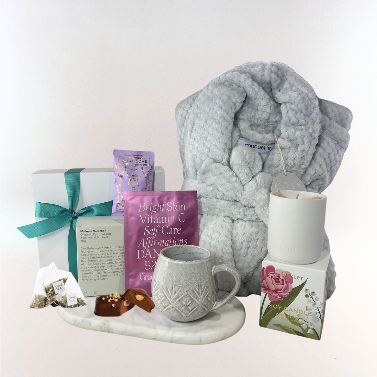 Cosy Self-Care Retreat gift box with a grey plush robe, organic wellness tea selection, textured stoneware mug, soy candle, chocolate pieces and face mask displayed on a marble tray