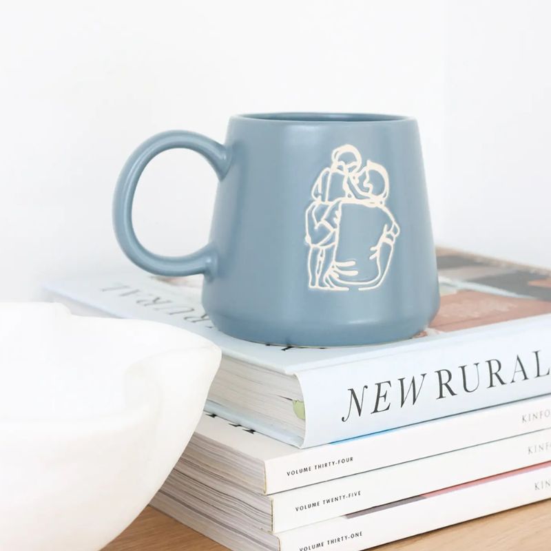 Minimalist ceramic mug with line art illustration of a couple embracing, styled on stacked books with modern home décor.