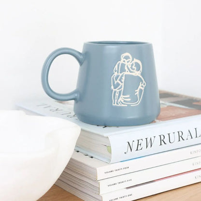 Minimalist ceramic mug with line art illustration of a couple embracing, styled on stacked books with modern home décor.