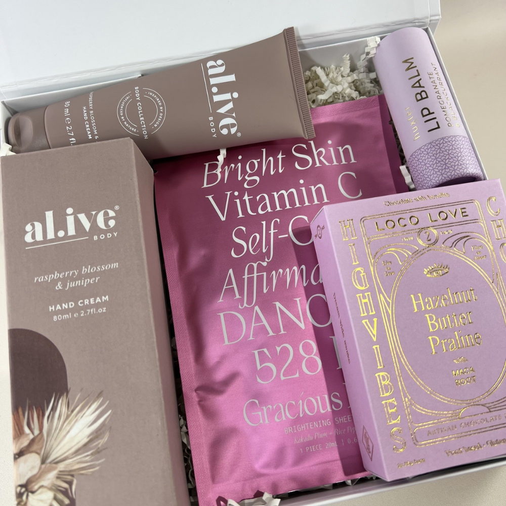The Luxe Lounge Rose Edition gift box open to reveal Al.ive Body Raspberry Blossom & Juniper hand cream, a Gracious Minds Bright Skin Vitamin C face mask, a pomegranate lip balm and Loco Love hazelnut butter praline chocolate, nestled in white paper shred