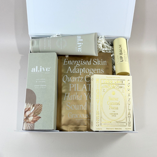 The Luxe Lounge gift box open to reveal Al.ive Body Sea Cotton & Coconut hand cream, a Lapcos face mask, Huxter lip balm and Loco Love butter caramel pecan chocolate, nestled in white paper shred