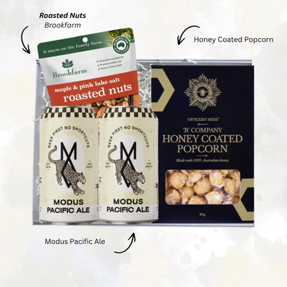 A gift-style product arrangement on a light off-white background: two Modus Pacific Ale cans in front of a black and gold 'Officiale Mess' honey coated popcorn tin, with a Brookfarm roasted nuts tag behind them. Arrows and labels point to the items, including 'Roasted Nuts Brookfarm' and 'Honey Coated Popcorn.