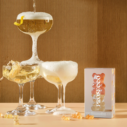 Vesper Bites Prosecco Bears gummies 100g in a blush pink box with a stack of orange and gold gummy bears printed on the front, surrounded by loose gummy bears and glasses.