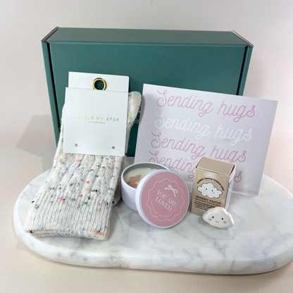 Sending Hugs gift box with speckled knit socks by Angels Whisper, a "You Are Loved" candle, a ceramic Worry Cloud keepsake and a "Sending Hugs" card on a marble tray, with dark green gift box behind