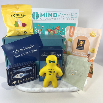 The Don't Stress Mate Box: a yellow Don't Stress Mate squishie wearing sunglasses, a Lemon + Olive Prize Catch bath salts tin, a Mind Waves crossword puzzle book, LOVE tea Sleep and Peppermint tea sachets, Byron Bay Cookies Macadamia Shortbread Bites, Funday Party Mix and a "Life is tough – but so are you" card, on a marble tray in front of a teal gift box