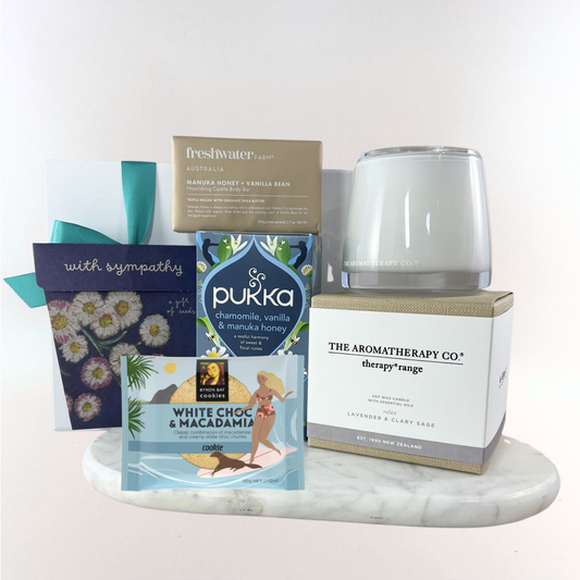 Thinking of You sympathy gift box with a calming lavender candle, forget-me-not seed packet, chamomile herbal tea, manuka honey soap bar, white chocolate macadamia cookie and a "With Sympathy" card on a marble tray