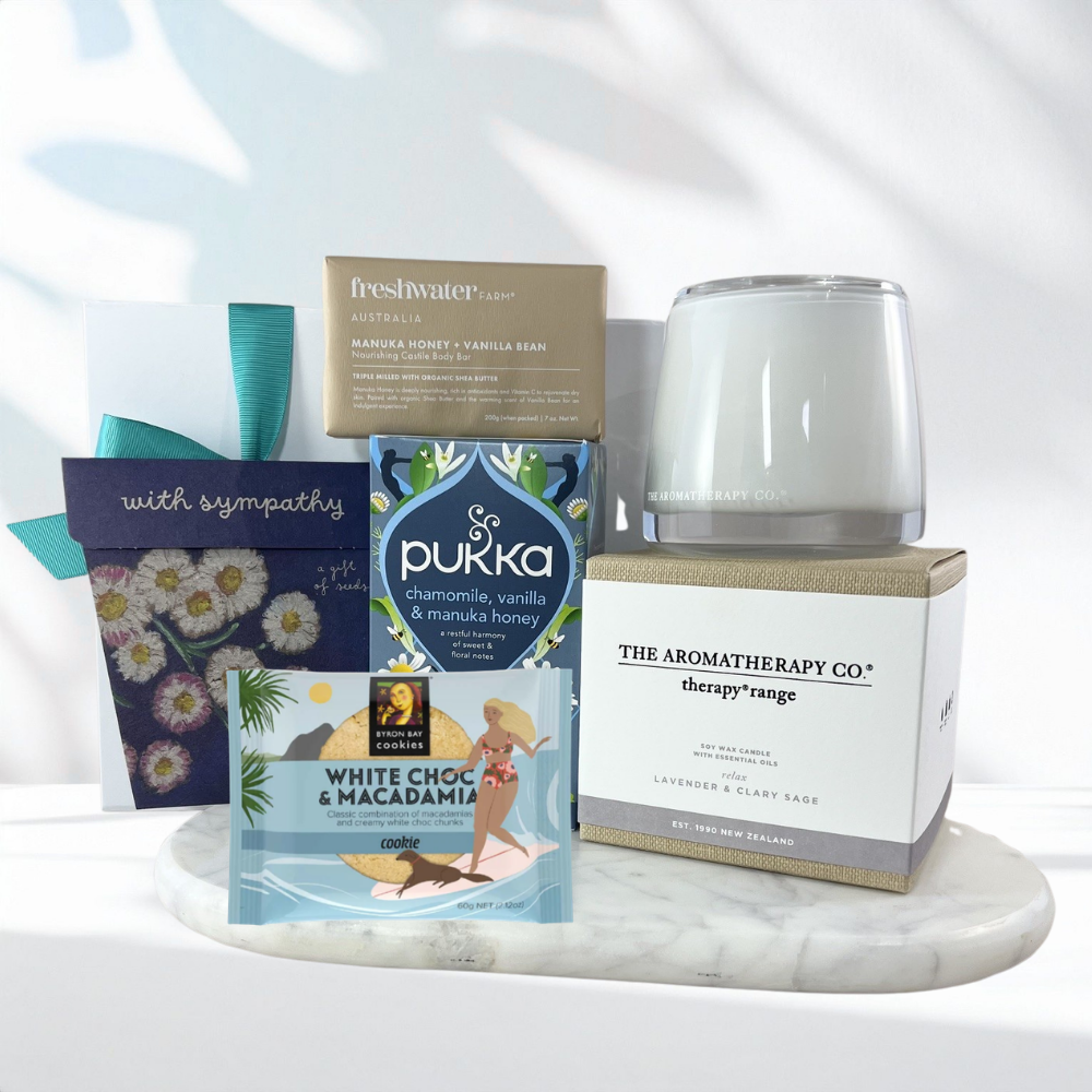 Thinking of You sympathy gift box with a calming lavender candle, forget-me-not seed packet, chamomile herbal tea, manuka honey soap bar, white chocolate macadamia cookie and a "With Sympathy" card on a marble tray