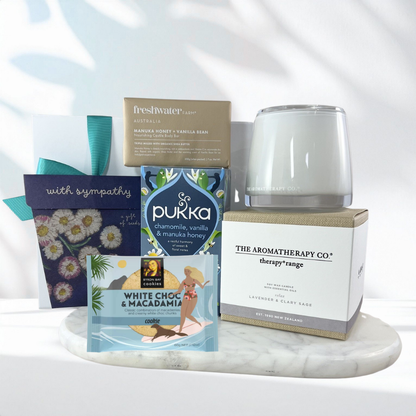 Thinking of You sympathy gift box with a calming lavender candle, forget-me-not seed packet, chamomile herbal tea, manuka honey soap bar, white chocolate macadamia cookie and a "With Sympathy" card on a marble tray