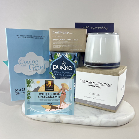 Thinking of You sympathy gift box with a calming lavender candle, forget-me-not seed packet, chamomile herbal tea, manuka honey soap bar, white chocolate macadamia cookie and a "With Sympathy" card on a marble tray. Includes Coping with Grief book.