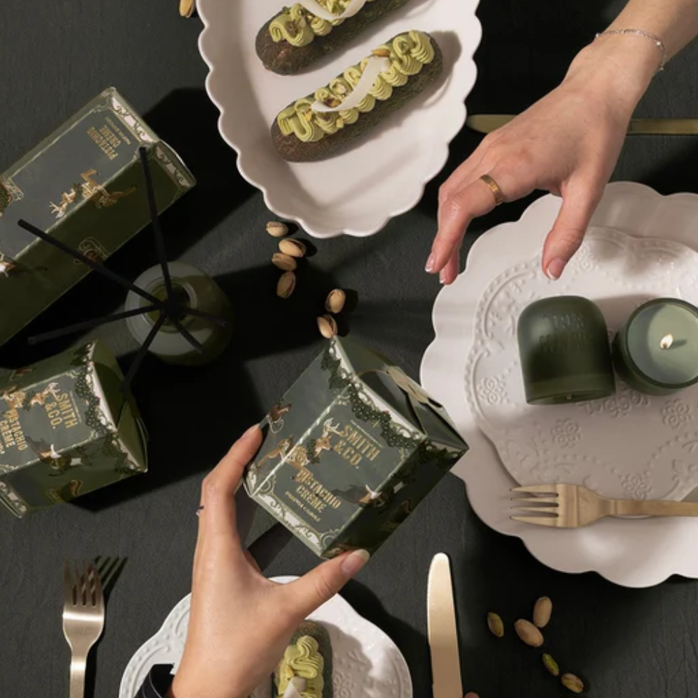 Hands holding a Smith & Co. Pistachio Crème candle gift box on a festive dinner table with pistachio desserts