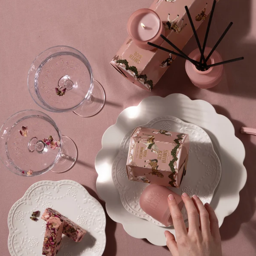 Hand holding a slice of nougat on a white plate beside a pink Smith & Co. reed diffuser and glassware