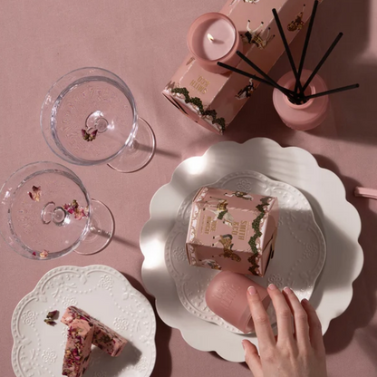 Hand holding a slice of nougat on a white plate beside a pink Smith & Co. reed diffuser and glassware
